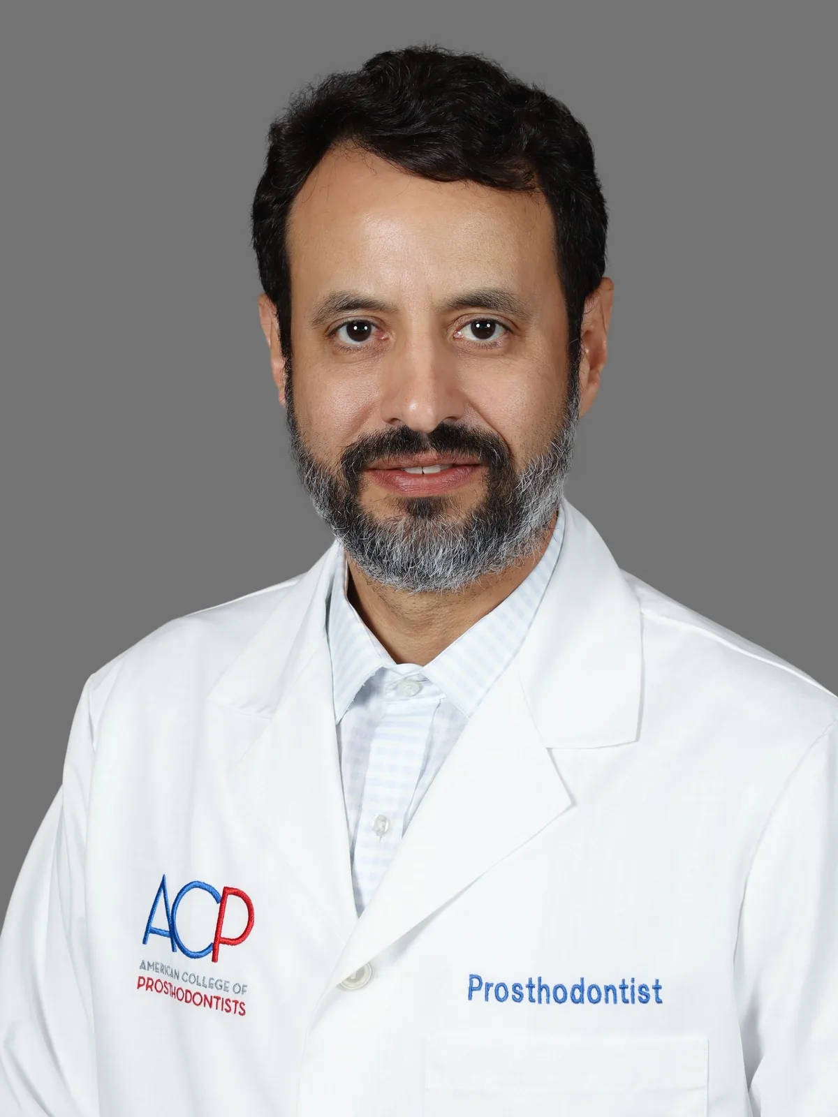 Studio portrait of Dr. Roy Rosado in a white coat embroidered with the ACP logo and Prosthodontist