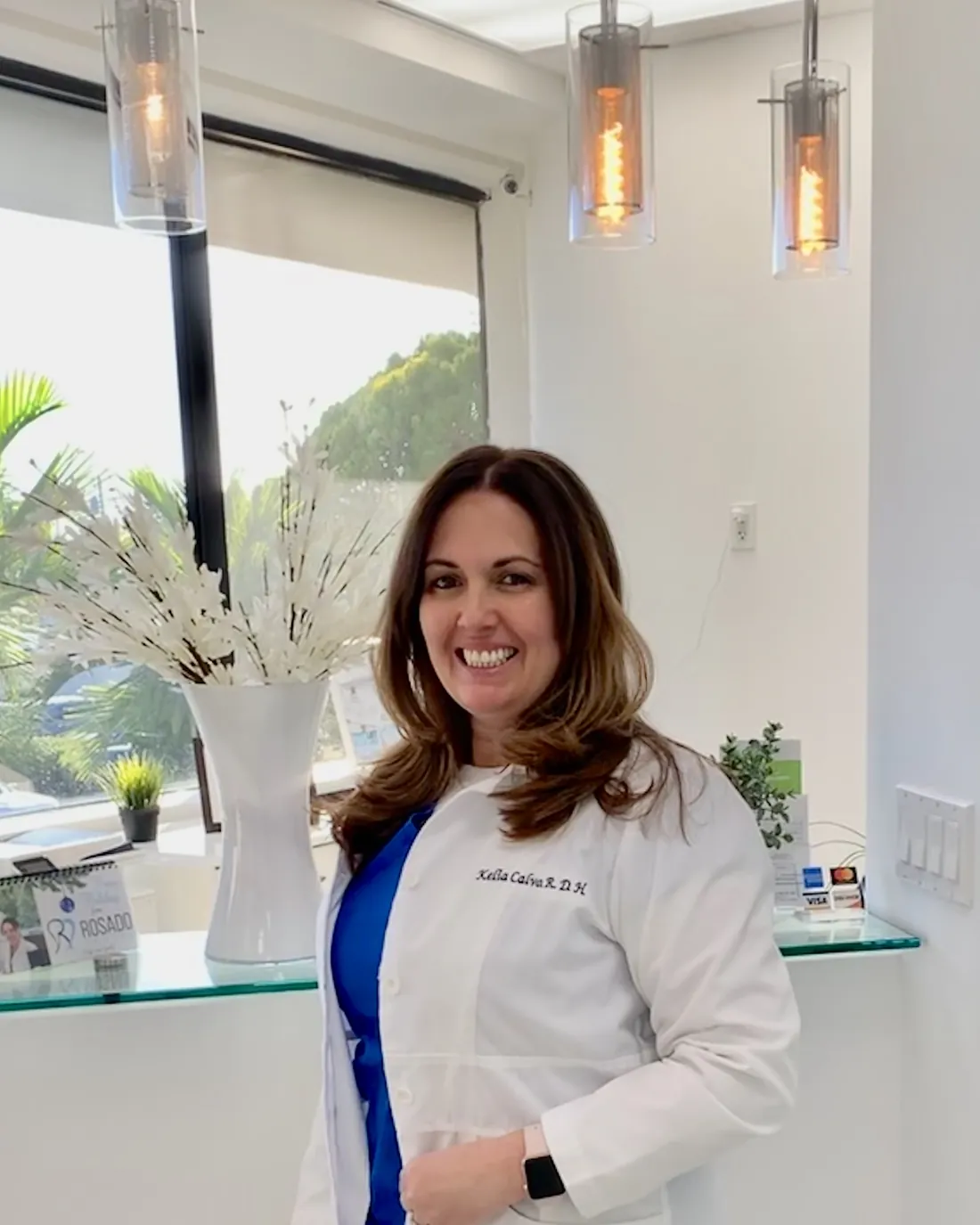 Kelia Calvo Rosado, RDH, smiling in a white lab coat at the Rosado Dental reception desk